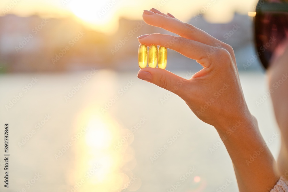 Yellow gel capsule vitamin d omega-3 in womans hand, sun sea background - obrazy, fototapety, plakaty 