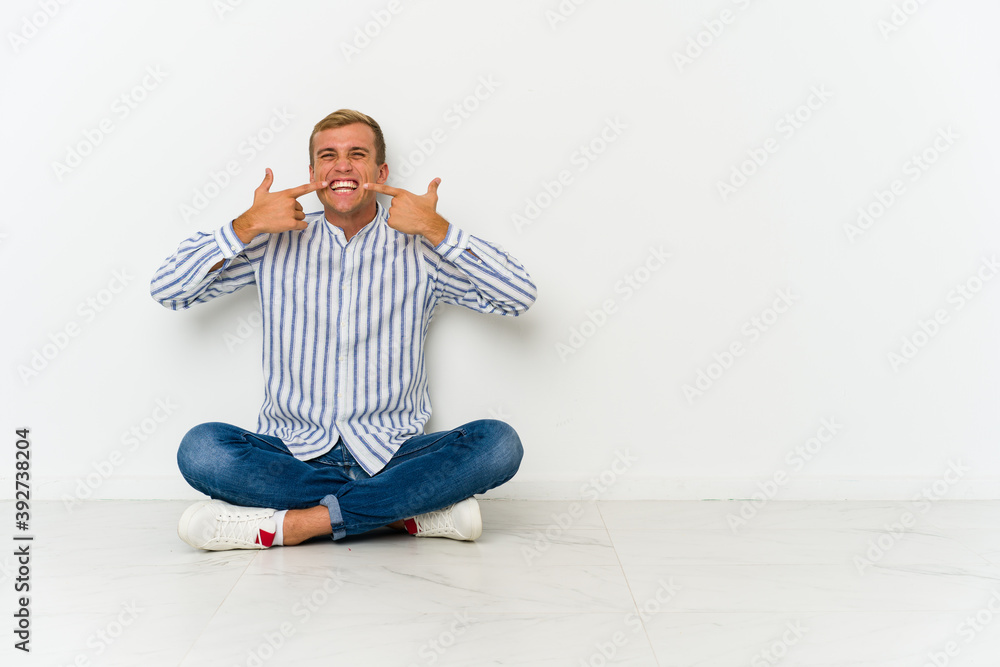 Young caucasian man sitting on the floor smiles, pointing fingers at mouth. Young caucasian man sitting on the floor smiles, pointing fingers at mouth.