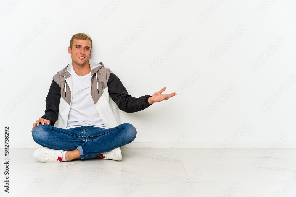 Young caucasian man sitting on the floor showing a welcome expression.
