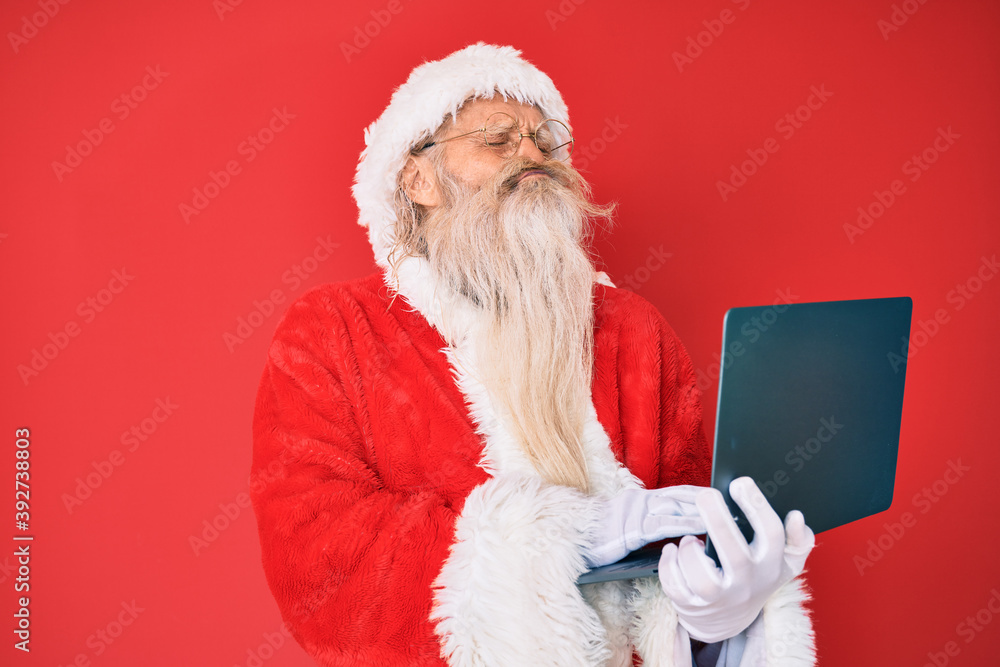 Naklejka premium Old senior man with grey hair and long beard wearing santa claus costume using laptop looking at the camera blowing a kiss being lovely and sexy. love expression.