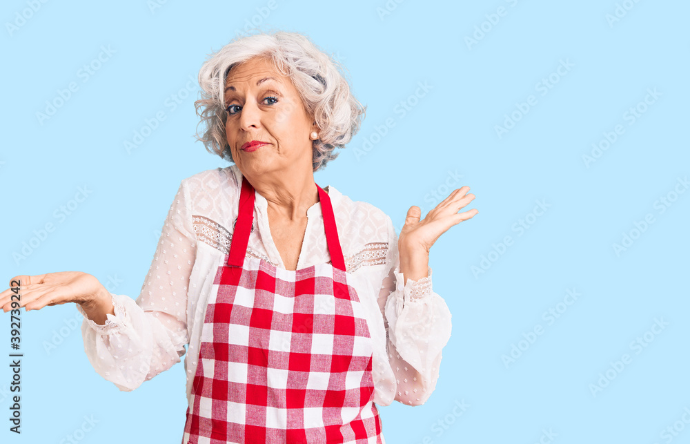 Senior grey-haired woman wearing apron clueless and confused expression with arms and hands raised. doubt concept. Senior grey-haired woman wearing apron clueless and confused expression with arms and hands raised. doubt concept.