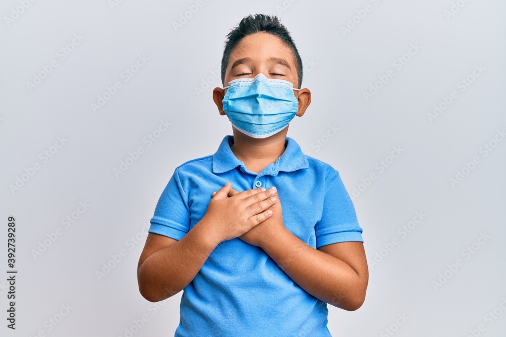 Little boy hispanic kid wearing medical mask smiling with hands on chest, eyes closed with grateful gesture on face. health concept.