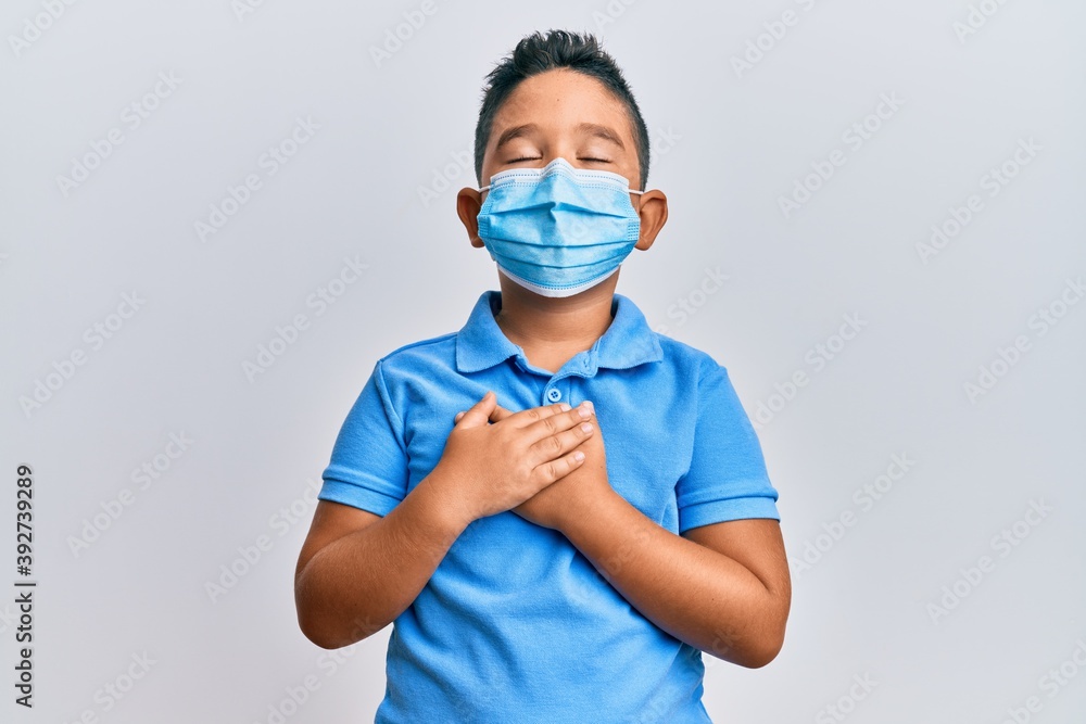 Little boy hispanic kid wearing medical mask smiling with hands on chest, eyes closed with grateful gesture on face. health concept. Little boy hispanic kid wearing medical mask smiling with hands on chest, eyes closed with grateful gesture on face. health concept.