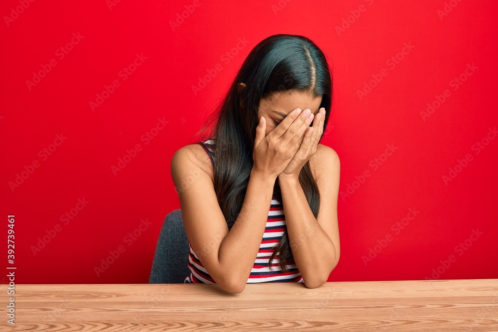Beautiful hispanic woman wearing casual clothes sitting on the table with sad expression covering face with hands while crying. depression concept. Beautiful hispanic woman wearing casual clothes sitting on the table with sad expression covering face with hands while crying. depression concept.