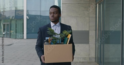 Coronacrisis and unemployment. Sad african american manager going outdoors along office, holding box of personal staff photo