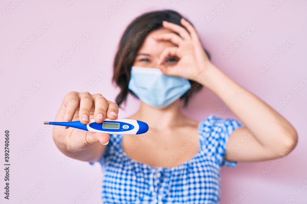 Young beautiful woman wearing medical mask holding holding thermometer smiling happy doing ok sign with hand on eye looking through fingers Young beautiful woman wearing medical mask holding holding thermometer smiling happy doing ok sign with hand on eye looking through fingers