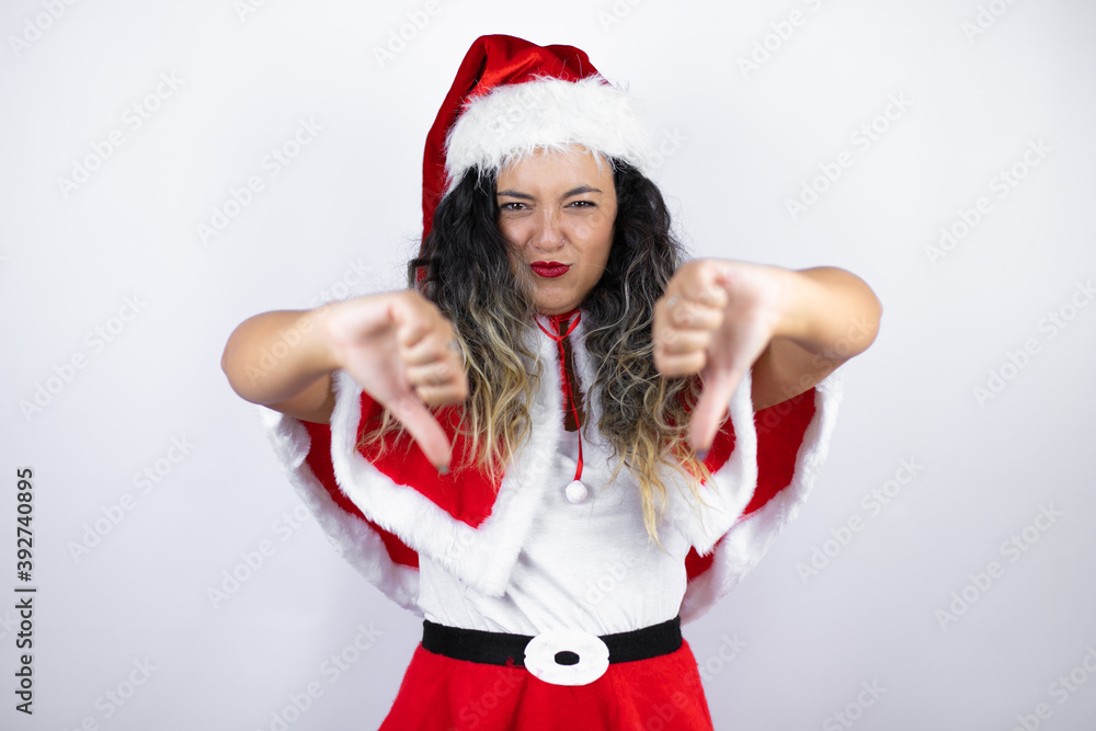 Young beautiful woman wearing a Santa Claus costume over white background with angry face, negative sign showing dislike with thumbs down Young beautiful woman wearing a Santa Claus costume over white background with angry face, negative sign showing dislike with thumbs down