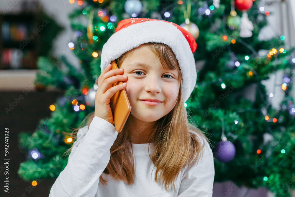 Online Christmas greetings. A close-up portrait of a cute girl in a New Year's hat with a mobile phone. The child uses gadgets to congratulate family and friends Online Christmas greetings. A close-up portrait of a cute girl in a New Year's hat with a mobile phone. The child uses gadgets to congratulate family and friends