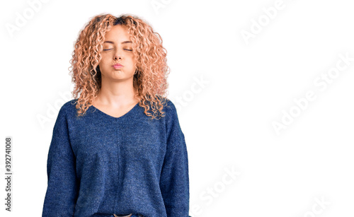 Young blonde woman with curly hair wearing casual winter sweater puffing cheeks with funny face. mouth inflated with air, crazy expression.