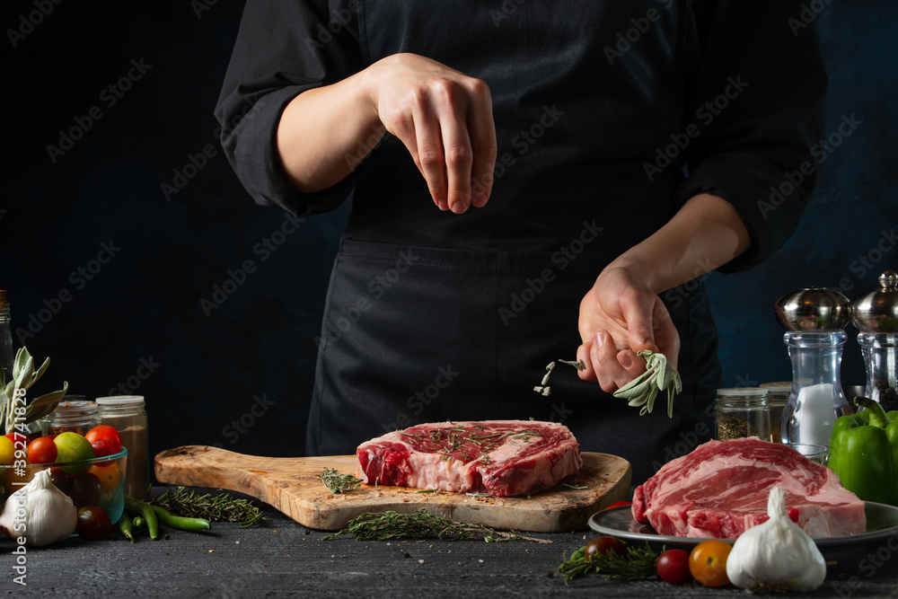 Professional chef pours some rosemary on raw steak on wooden chopped board. Backstage of preparing grilled pork meat at restaurant kitchen on dark blue background. Frozen motion.