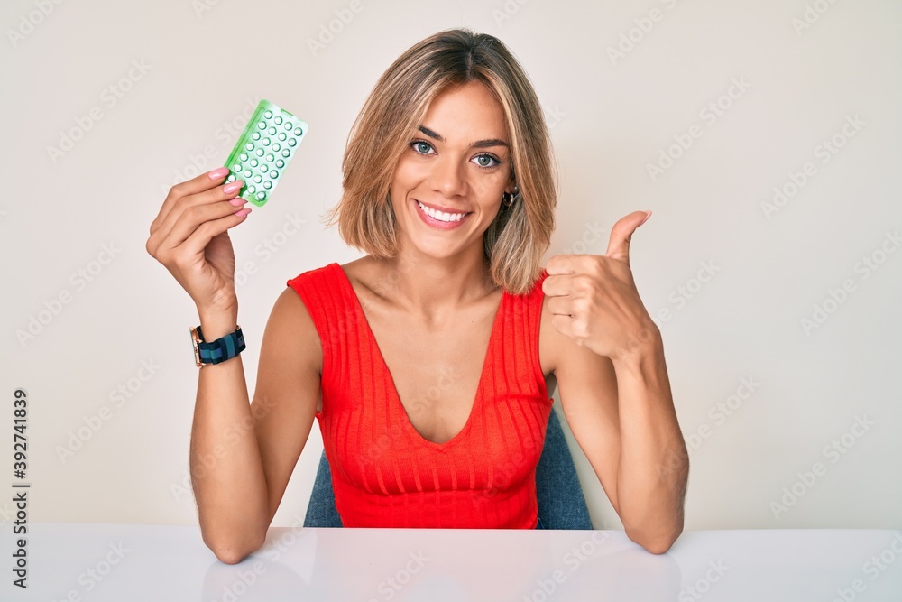 Beautiful caucasian woman holding birth control pills smiling happy and positive, thumb up doing excellent and approval sign Beautiful caucasian woman holding birth control pills smiling happy and positive, thumb up doing excellent and approval sign