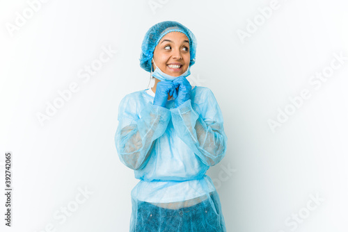 Young surgeon latin woman isolated on white background keeps hands under chin, is looking happily aside. © Asier