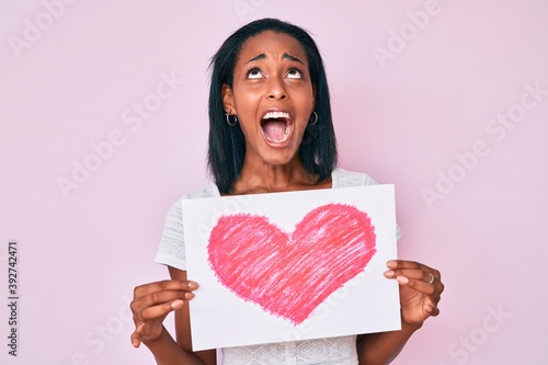 Young african american woman holding heart draw angry and mad screaming frustrated and furious  shouting with anger looking up.