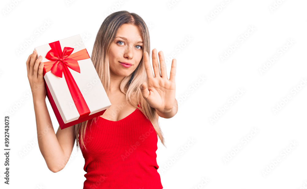 Young beautiful blonde woman holding gift with open hand doing stop sign with serious and confident expression, defense gesture Young beautiful blonde woman holding gift with open hand doing stop sign with serious and confident expression, defense gesture