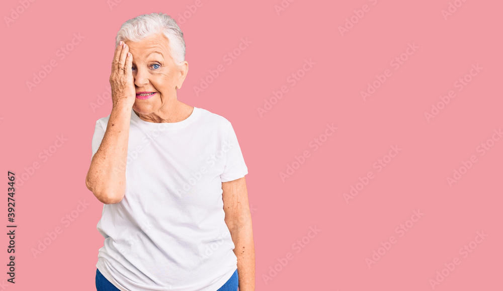 Senior beautiful woman with blue eyes and grey hair wearing casual white tshirt covering one eye with hand, confident smile on face and surprise emotion. Senior beautiful woman with blue eyes and grey hair wearing casual white tshirt covering one eye with hand, confident smile on face and surprise emotion.