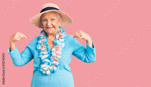 Senior beautiful woman with blue eyes and grey hair wearing summer hat and hawaiian lei looking confident with smile on face, pointing oneself with fingers proud and happy. © Krakenimages.com