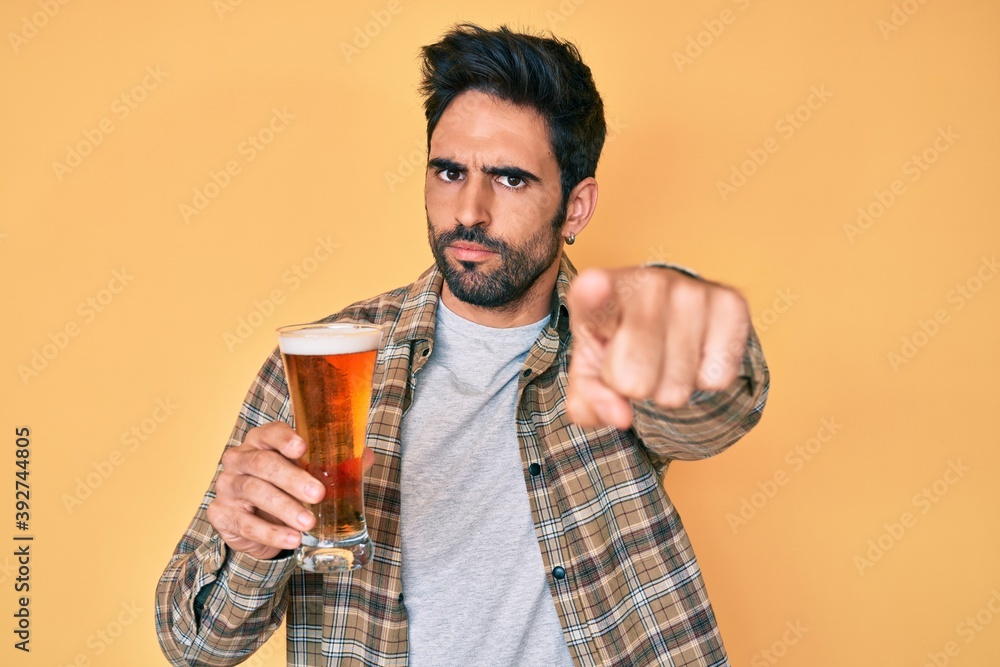 Handsome hispanic man with beard drinking a pint of beer pointing with finger to the camera and to you, confident gesture looking serious Handsome hispanic man with beard drinking a pint of beer pointing with finger to the camera and to you, confident gesture looking serious
