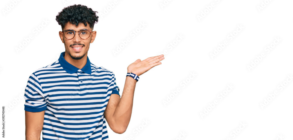 Young arab man wearing casual clothes and glasses smiling cheerful presenting and pointing with palm of hand looking at the camera. Young arab man wearing casual clothes and glasses smiling cheerful presenting and pointing with palm of hand looking at the camera.