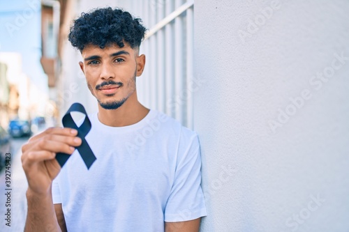 Young arab man with serious expression holding mouring black ribbon leaning on the wall. photo