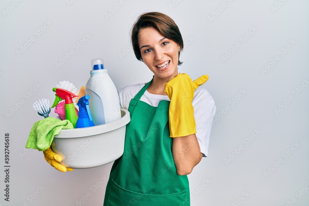 Young brunette woman with short hair wearing apron holding cleaning products smiling with happy face looking and pointing to the side with thumb up. Young brunette woman with short hair wearing apron holding cleaning products smiling with happy face looking and pointing to the side with thumb up.