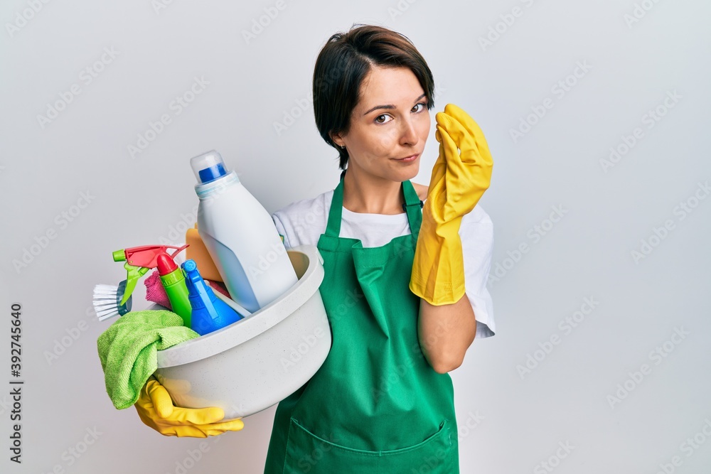 Young brunette woman with short hair wearing apron holding cleaning products doing italian gesture with hand and fingers confident expression Young brunette woman with short hair wearing apron holding cleaning products doing italian gesture with hand and fingers confident expression