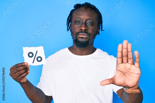 Young african american man with braids holding percentage symbol paper with open hand doing stop sign with serious and confident expression, defense gesture © Krakenimages.com