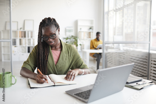 Front view portrait of contemporary African-American woman writing in planner while working at desk in white office interior, copy space