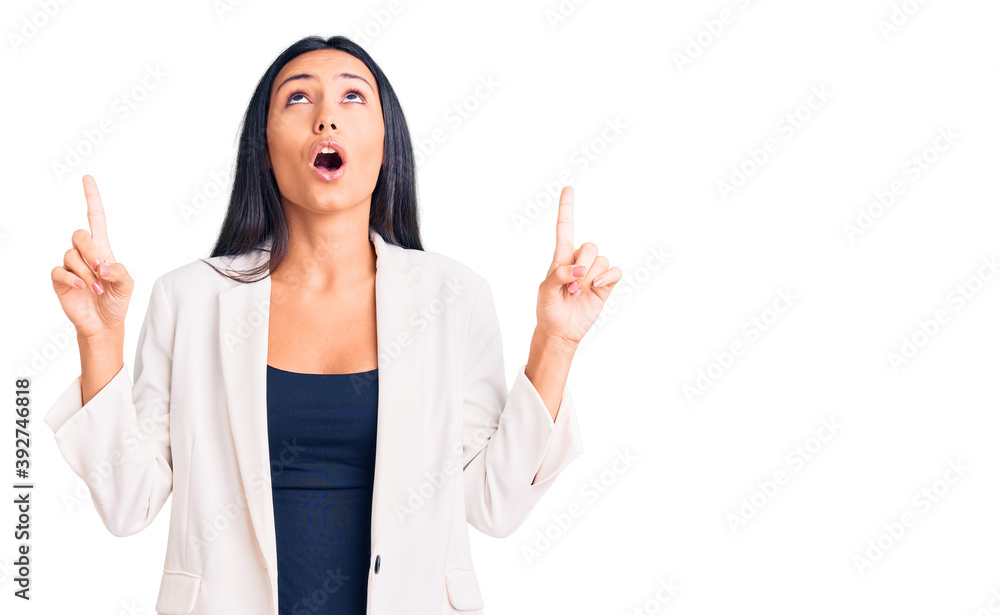 Young beautiful latin girl wearing business clothes amazed and surprised looking up and pointing with fingers and raised arms. Young beautiful latin girl wearing business clothes amazed and surprised looking up and pointing with fingers and raised arms.