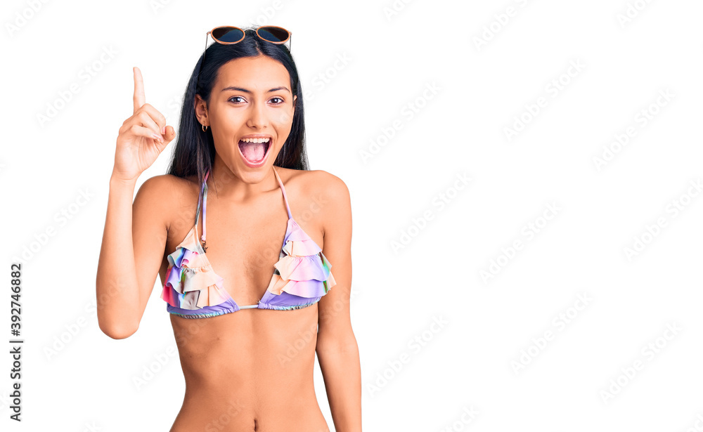 Obraz premium Young beautiful latin girl wearing bikini and sunglasses pointing finger up with successful idea. exited and happy. number one.