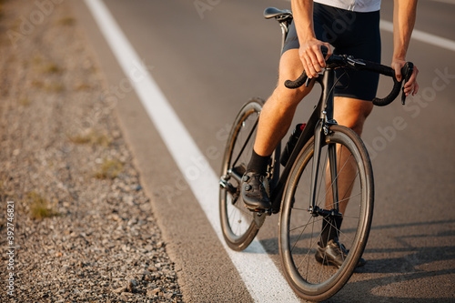 Young sportsman in activewear and sneakers sitting on black bike on paved road. Close up of strong male legs. Regular training on fresh air.