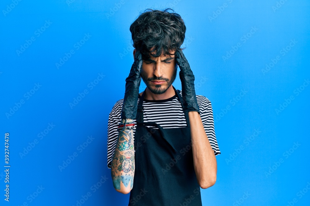 Young hispanic man wearing barber apron suffering from headache desperate and stressed because pain and migraine. hands on head.