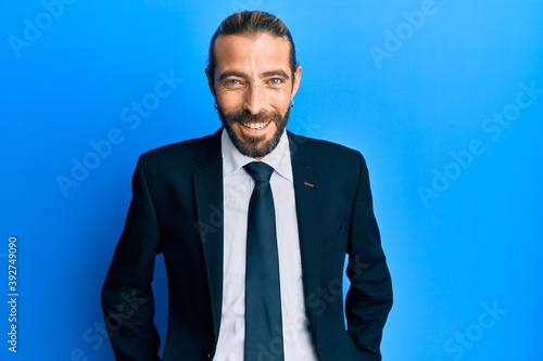 Attractive man with long hair and beard wearing business suit and tie looking positive and happy standing and smiling with a confident smile showing teeth