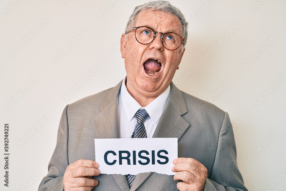 Senior grey-haired man wearing business suit holding crisis paper angry and mad screaming frustrated and furious, shouting with anger looking up. Senior grey-haired man wearing business suit holding crisis paper angry and mad screaming frustrated and furious, shouting with anger looking up.