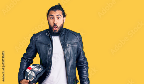 Young arab man holding motorcycle helmet scared and amazed with open mouth for surprise, disbelief face