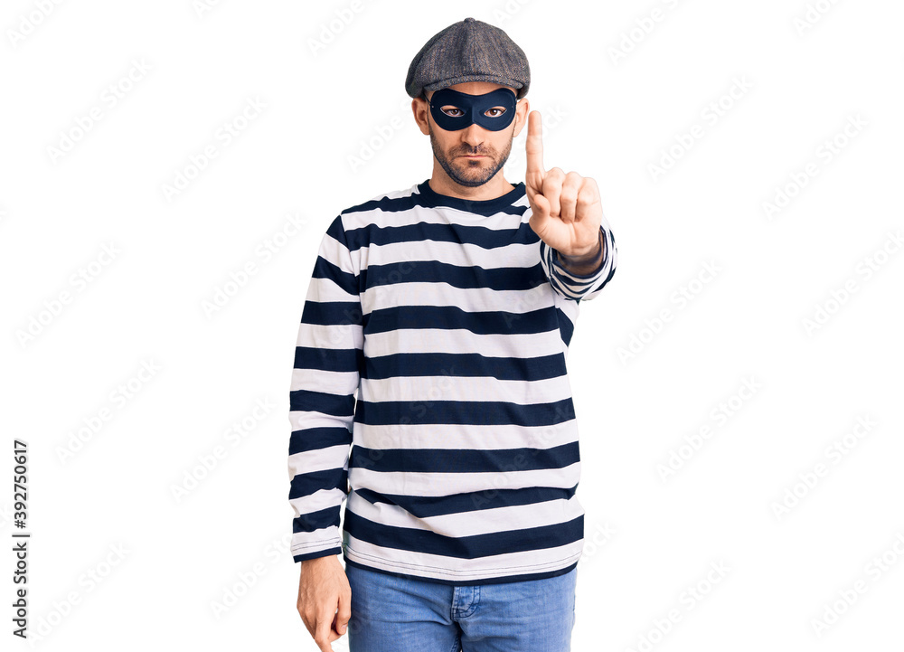 Young handsome man wearing burglar mask pointing with finger up and angry expression, showing no gesture Young handsome man wearing burglar mask pointing with finger up and angry expression, showing no gesture
