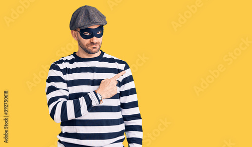 Young handsome man wearing burglar mask pointing with hand finger to the side showing advertisement, serious and calm face