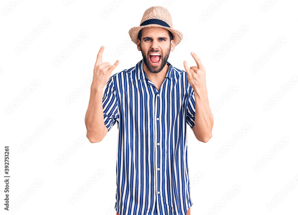 Young handsome man with beard wearing summer hat and striped shirt shouting with crazy expression doing rock symbol with hands up. music star. heavy concept. Young handsome man with beard wearing summer hat and striped shirt shouting with crazy expression doing rock symbol with hands up. music star. heavy concept.