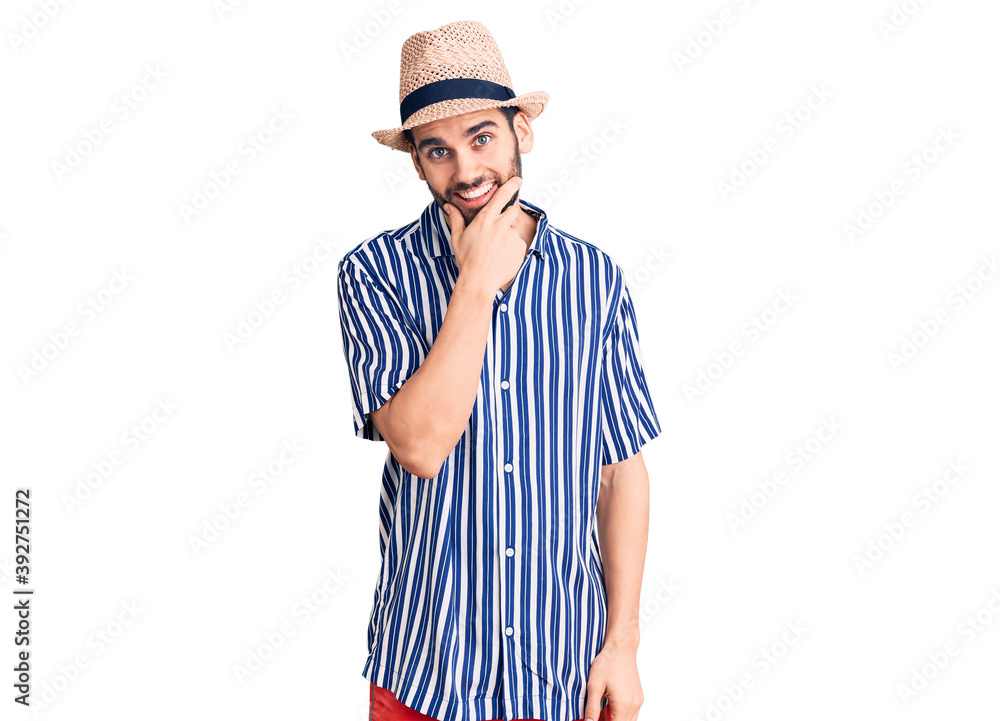 Young handsome man with beard wearing summer hat and striped shirt looking confident at the camera smiling with crossed arms and hand raised on chin. thinking positive. Young handsome man with beard wearing summer hat and striped shirt looking confident at the camera smiling with crossed arms and hand raised on chin. thinking positive.