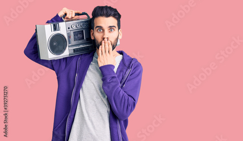 Young handsome man with beard listening to music using vintage boombox covering mouth with hand, shocked and afraid for mistake. surprised expression