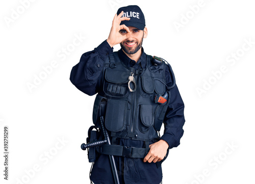 Young handsome man with beard wearing police uniform doing ok gesture with hand smiling, eye looking through fingers with happy face.