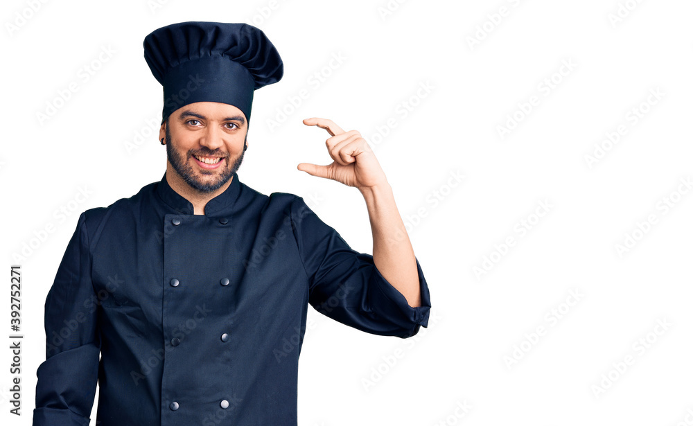 Young hispanic man wearing cooker uniform smiling and confident gesturing with hand doing small size sign with fingers looking and the camera. measure concept. Young hispanic man wearing cooker uniform smiling and confident gesturing with hand doing small size sign with fingers looking and the camera. measure concept.