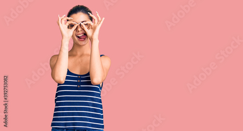 Young beautiful woman wearing casual clothes and glasses doing ok gesture like binoculars sticking tongue out, eyes looking through fingers. crazy expression.