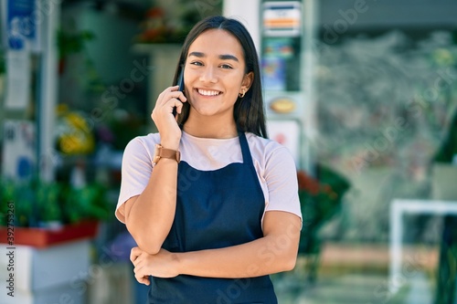 Young latin shopkeeper girl smiling happy talking on the smartphone at florist.