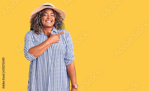 Young african american plus size woman wearing summer hat cheerful with a smile of face pointing with hand and finger up to the side with happy and natural expression on face