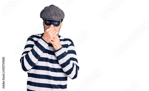 Middle age handsome man wearing burglar mask smelling something stinky and disgusting, intolerable smell, holding breath with fingers on nose. bad smell