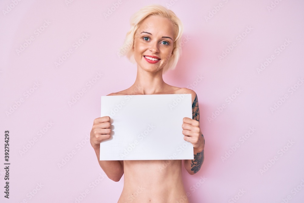 Young blonde woman with tattoo standing shirtless holding empty blank banner looking positive and happy standing and smiling with a confident smile showing teeth Young blonde woman with tattoo standing shirtless holding empty blank banner looking positive and happy standing and smiling with a confident smile showing teeth