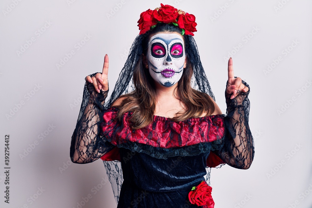Young woman wearing day of the dead costume over white smiling amazed and surprised and pointing up with fingers and raised arms. Young woman wearing day of the dead costume over white smiling amazed and surprised and pointing up with fingers and raised arms.