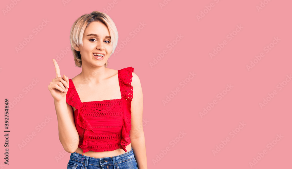 Young beautiful blonde woman wearing casual clothes showing and pointing up with finger number one while smiling confident and happy. Young beautiful blonde woman wearing casual clothes showing and pointing up with finger number one while smiling confident and happy.