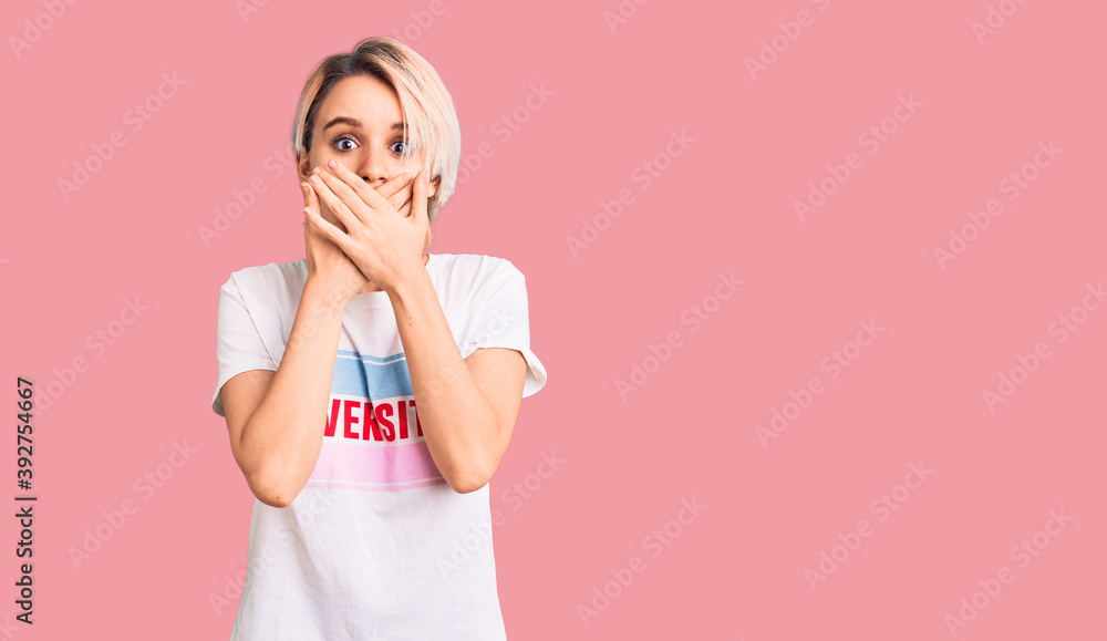 Young beautiful blonde woman wearing t shirt with diversity word message shocked covering mouth with hands for mistake. secret concept. Young beautiful blonde woman wearing t shirt with diversity word message shocked covering mouth with hands for mistake. secret concept.