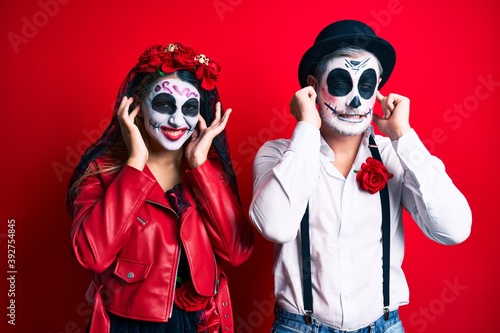 Couple wearing day of the dead costume over red covering ears with fingers with annoyed expression for the noise of loud music. deaf concept.